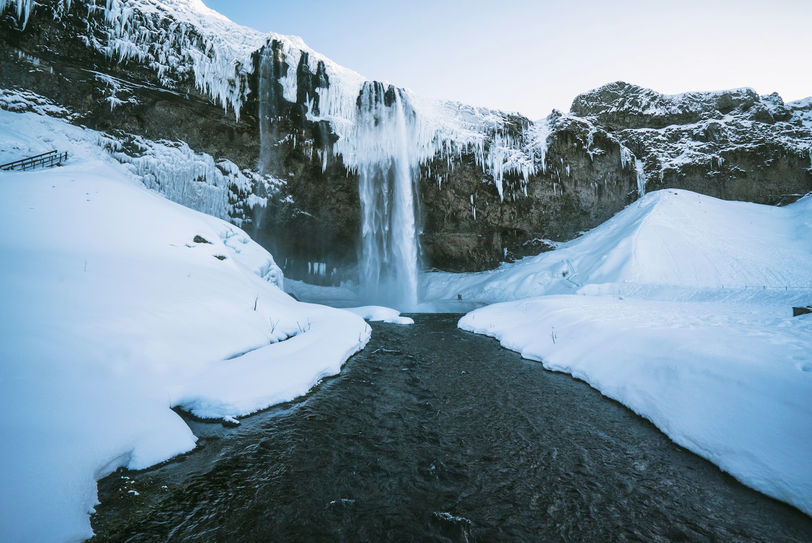 A large Icelandic waterfall falling from an icicle-fringed cliff, surrounded by snowy banks and a dark stream