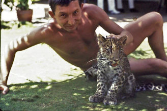 A man is sitting on the grass with a young leopard beside him. The setting appears outdoors with sunlight filtering through leaves, casting shadows. The man is shirtless, relaxed, and smiling, while the leopard cub seems calmly alert.