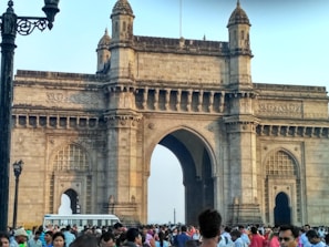 An ornate stone arch, known as the Gateway of India, stands prominently against a clear sky. This historic monument features intricate latticework and carvings. A large crowd of people gathers in front of the structure, with several individuals taking photos. A section of a bus is visible on the left side, emphasizing the tourist nature of the location.