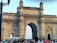 An ornate stone arch, known as the Gateway of India, stands prominently against a clear sky. This historic monument features intricate latticework and carvings. A large crowd of people gathers in front of the structure, with several individuals taking photos. A section of a bus is visible on the left side, emphasizing the tourist nature of the location.