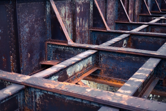Rusted steel beams form a grid-like structure with visible signs of weathering and oxidation. The metal surfaces are covered with patches of rust, creating a textured appearance. The beams intersect at right angles, creating a sense of industrial design and architecture.