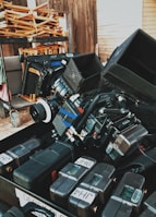 A collection of professional camera equipment and accessories are arranged in a dense, organized manner. The scene includes stacked electronic gear, camera lenses, and various attachments, suggesting a film set or prepping area. In the background, folded wooden chairs are placed on a rack, and wooden walls set a rustic tone.