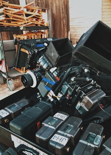 A collection of professional camera equipment and accessories are arranged in a dense, organized manner. The scene includes stacked electronic gear, camera lenses, and various attachments, suggesting a film set or prepping area. In the background, folded wooden chairs are placed on a rack, and wooden walls set a rustic tone.