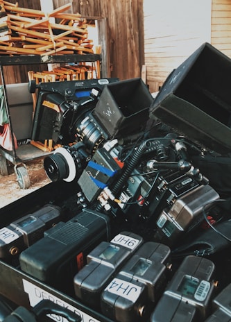 A collection of professional camera equipment and accessories are arranged in a dense, organized manner. The scene includes stacked electronic gear, camera lenses, and various attachments, suggesting a film set or prepping area. In the background, folded wooden chairs are placed on a rack, and wooden walls set a rustic tone.