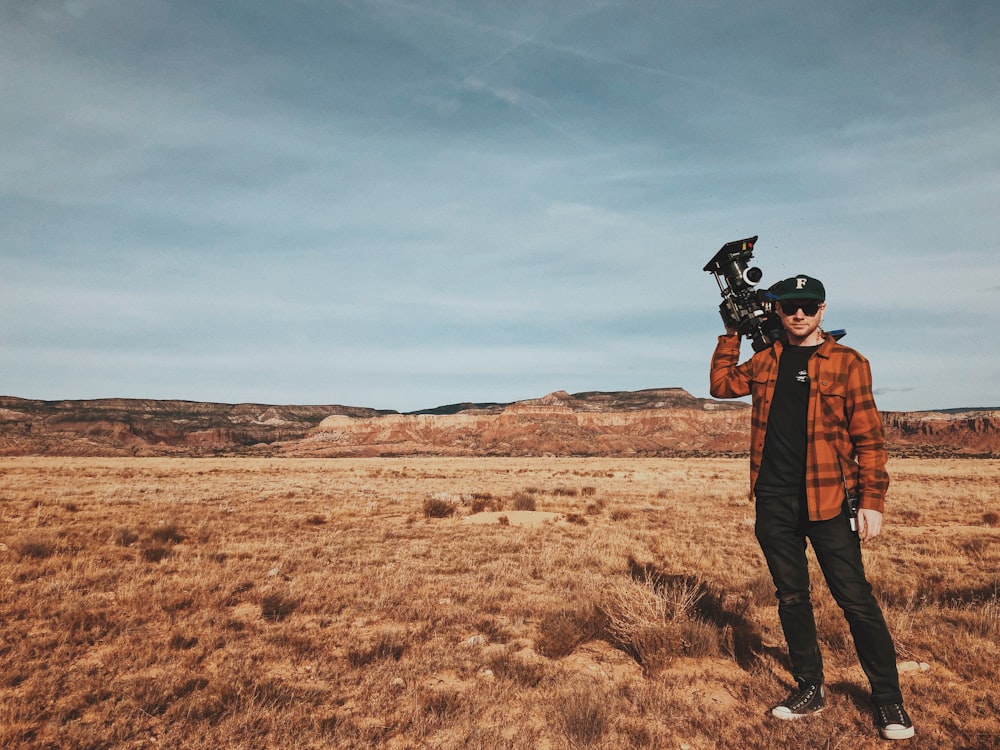 Homme debout sur un champ brun portant une caméra d’enregistrement noire pendant la journée