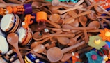 A collection of wooden spoons scattered with vibrant wooden toys and tambourines intermixed. The toys are brightly colored, featuring elements like an orange animal, drumheads, and decorative flowers.