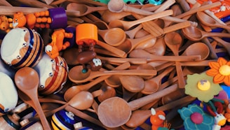 A collection of wooden spoons scattered with vibrant wooden toys and tambourines intermixed. The toys are brightly colored, featuring elements like an orange animal, drumheads, and decorative flowers.
