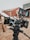 A professional video camera setup is positioned outside, with a wooden building in the background. The camera is mounted on a tripod and has several attachments, including a monitor displaying a scene of two people standing in front of a vintage car. The environment suggests a film set or production area with an old-western theme.