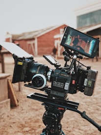 A professional video camera setup is positioned outside, with a wooden building in the background. The camera is mounted on a tripod and has several attachments, including a monitor displaying a scene of two people standing in front of a vintage car. The environment suggests a film set or production area with an old-western theme.
