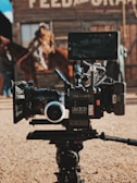 A professional film camera is set up in the foreground, with a rustic, wooden building and an out-of-focus person riding a horse in the background. The camera is detailed with various attachments, including a large lens and a monitor.