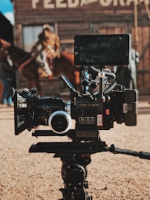A professional film camera is set up in the foreground, with a rustic, wooden building and an out-of-focus person riding a horse in the background. The camera is detailed with various attachments, including a large lens and a monitor.