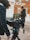 A filmmaker is operating a professional camera mounted on a tripod, capturing a scene from a film set that resembles a Western town. The camera's monitor displays an image of a person dressed in a period costume, suggesting the filming of a historical or Western-themed production. The person near the camera is dressed warmly, indicating cool weather.