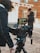 A filmmaker is operating a professional camera mounted on a tripod, capturing a scene from a film set that resembles a Western town. The camera's monitor displays an image of a person dressed in a period costume, suggesting the filming of a historical or Western-themed production. The person near the camera is dressed warmly, indicating cool weather.