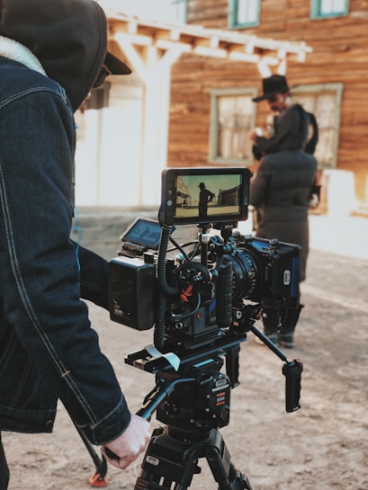 A filmmaker is operating a professional camera mounted on a tripod, capturing a scene from a film set that resembles a Western town. The camera's monitor displays an image of a person dressed in a period costume, suggesting the filming of a historical or Western-themed production. The person near the camera is dressed warmly, indicating cool weather.