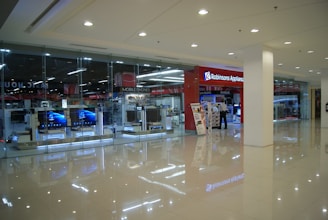 Inside a shopping mall, there is a store with a glass facade displaying electronic products such as televisions. The sign 'Robinsons Appliance' indicates the store's name. Nearby, a person stands next to promotional displays, and the interior is well-lit with polished floors reflecting the lighting.