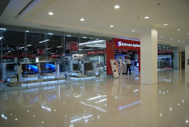 Inside a shopping mall, there is a store with a glass facade displaying electronic products such as televisions. The sign 'Robinsons Appliance' indicates the store's name. Nearby, a person stands next to promotional displays, and the interior is well-lit with polished floors reflecting the lighting.