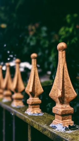 A row of ornate, golden metallic fence posts with a pointed design, set against a lush, dark green blurred background. The fence displays a patina of age, enhancing the rustic appearance.