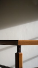 A wooden table corner is captured with sunlight casting soft, diagonal shadows on a light gray wall. The table features a simple design with a focus on clean lines and natural wood grain.