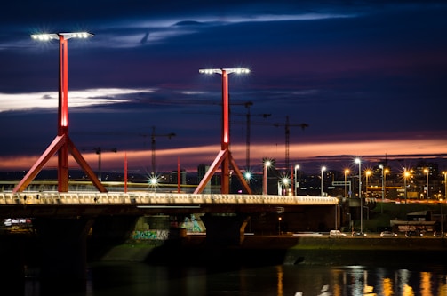 A well-lit bridge spans a river under a dramatic, darkening sky. The bridge features modern, red structural beams and multiple streetlamps illuminating its length. In the background, construction cranes are silhouetted against a vibrant sunset with hues of orange and purple.