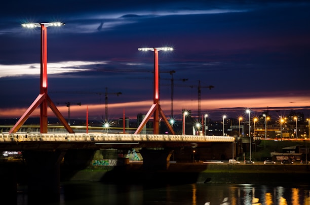 A well-lit bridge spans a river under a dramatic, darkening sky. The bridge features modern, red structural beams and multiple streetlamps illuminating its length. In the background, construction cranes are silhouetted against a vibrant sunset with hues of orange and purple.