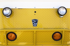 A close-up view of the front of a yellow industrial vehicle with two round headlights and two orange reflectors. The surface features a diamond plate pattern and a logo that reads 'Taylor Dunn' in the center.