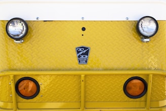 A close-up view of the front of a yellow industrial vehicle with two round headlights and two orange reflectors. The surface features a diamond plate pattern and a logo that reads 'Taylor Dunn' in the center.