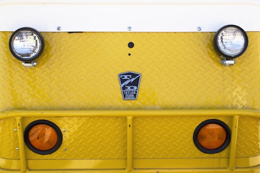 A close-up view of the front of a yellow industrial vehicle with two round headlights and two orange reflectors. The surface features a diamond plate pattern and a logo that reads 'Taylor Dunn' in the center.