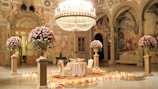 An opulent setting features a grand chandelier hanging above an elegantly decorated table for two, surrounded by large vases overflowing with pink roses. The room is adorned with detailed frescoes and classic architectural elements, and the floor is lined with candles and rose petals, creating a romantic ambiance.