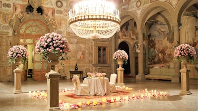 An opulent setting features a grand chandelier hanging above an elegantly decorated table for two, surrounded by large vases overflowing with pink roses. The room is adorned with detailed frescoes and classic architectural elements, and the floor is lined with candles and rose petals, creating a romantic ambiance.