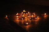 Numerous small clay lamps with flickering flames are arranged on a dark surface, casting warm orange and yellow hues. The lighting creates a serene and intimate ambiance, highlighting the lamps against the dim background.