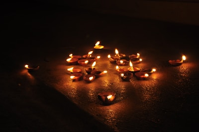 Numerous small clay lamps with flickering flames are arranged on a dark surface, casting warm orange and yellow hues. The lighting creates a serene and intimate ambiance, highlighting the lamps against the dim background.