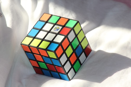 A multicolored Rubik's Cube with a sticker in its center is positioned on a white fabric surface. The cube displays a mix of bright colors, including red, blue, green, yellow, orange, and white, with sunlight casting shadows around it.