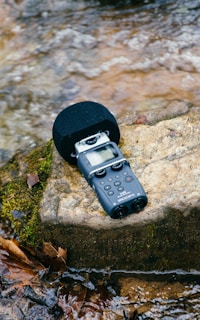 A portable audio recorder rests on a moss-covered rock beside a gently flowing stream. The device has a microphone and various controls and buttons on its body. Surrounding the rock are fallen leaves, adding a touch of nature to the scene.