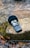 A portable audio recorder rests on a moss-covered rock beside a gently flowing stream. The device has a microphone and various controls and buttons on its body. Surrounding the rock are fallen leaves, adding a touch of nature to the scene.