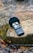 A portable audio recorder rests on a moss-covered rock beside a gently flowing stream. The device has a microphone and various controls and buttons on its body. Surrounding the rock are fallen leaves, adding a touch of nature to the scene.