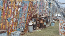 A row of colorful, intricately painted wooden panels leaning against a wall, with a variety of rusted and weathered paint cans and buckets placed in front. The grass-covered ground indicates an outdoor setting.
