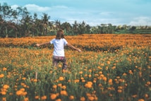 A person stands with outstretched arms in a vibrant field full of orange marigold flowers, surrounded by lush greenery and under a partly cloudy sky. The atmosphere is serene and expansive, suggesting freedom and joy.
