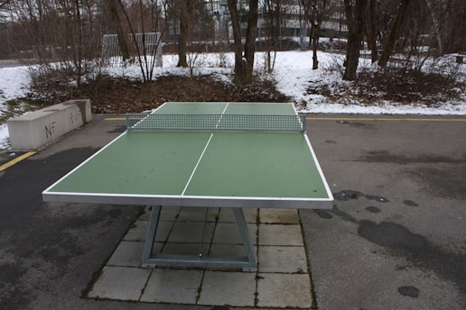 A green outdoor ping pong table with a metal net is set up on a concrete surface. The surrounding area shows a winter scene with patches of snow on the ground and bare trees. Nearby, a concrete bench is positioned next to a pathway, and a building is visible in the background.