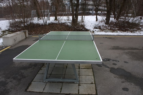 A green outdoor ping pong table with a metal net is set up on a concrete surface. The surrounding area shows a winter scene with patches of snow on the ground and bare trees. Nearby, a concrete bench is positioned next to a pathway, and a building is visible in the background.