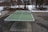 A green outdoor ping pong table with a metal net is set up on a concrete surface. The surrounding area shows a winter scene with patches of snow on the ground and bare trees. Nearby, a concrete bench is positioned next to a pathway, and a building is visible in the background.