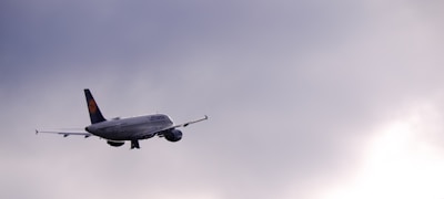 A commercial airplane with Lufthansa branding is in flight against a backdrop of a cloudy sky. The aircraft is flying towards the right side of the picture and appears to be cruising at a high altitude. The sky is mostly overcast with a light gradient from darker to lighter shades of grey.