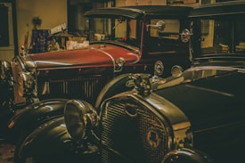 Two vintage cars are parked in a dimly lit garage. The cars appear to be classic models from the early 20th century, featuring detailed chrome accents and large headlights. The surrounding environment includes various tools and boxes, suggesting a workshop setting.