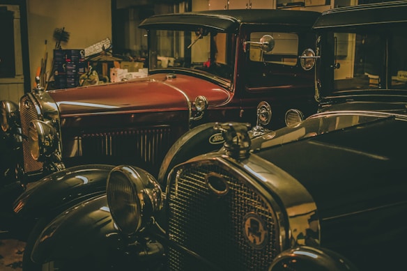 Two vintage cars are parked in a dimly lit garage. The cars appear to be classic models from the early 20th century, featuring detailed chrome accents and large headlights. The surrounding environment includes various tools and boxes, suggesting a workshop setting.