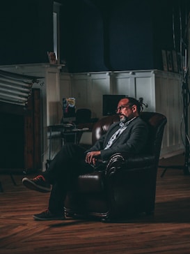 A man sits comfortably in a dark, leather armchair within a dimly lit room. The ambiance is enhanced by the dark walls and wooden flooring. Light from an unseen source casts shadows, creating a dramatic and contemplative atmosphere. In the background, there is a desk with a computer and various photography equipment, suggesting a studio or workspace environment.