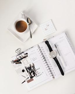 analog watch on top of book beside white ceramic teacup with teaspoon