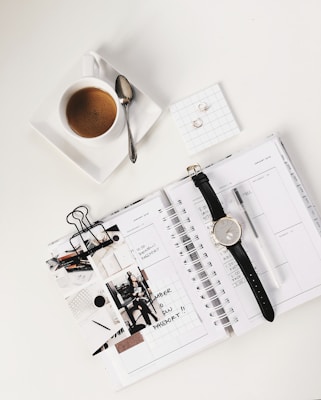 An overhead view of a neatly arranged workspace featuring a cup of coffee with a spoon on a saucer, a wristwatch on a planner, notepads, paper clips, a pen, some photographs, and a pair of earrings on a grid-marked square card.