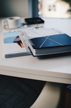 An open spiral notebook with a black cover featuring intersecting lines rests on top of a magazine on a white table. In the blurred background, there’s a white cup and a pair of keys placed on a dark surface.