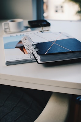 An open spiral notebook with a black cover featuring intersecting lines rests on top of a magazine on a white table. In the blurred background, there&rsquo;s a white cup and a pair of keys placed on a dark surface.