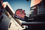 A neon sign with the words 'Twist Music Bar' and a star symbol is prominently displayed on an outdoor wall, indicating a music bar. There is also a smaller 'Pepsi' sign beneath it. The surroundings include buildings with balconies, a lot of electrical wires, and a clear sky during daylight.