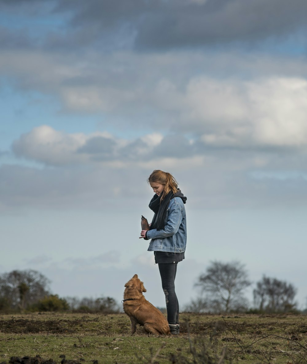 woman in blue denim jacket standing beside short-coated brown dog